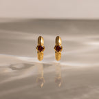 The Ribbed Garnet Huggies, gold hoop earrings with red gemstones, stand upright on a softly lit reflective surface—making them a perfect meaningful gift.