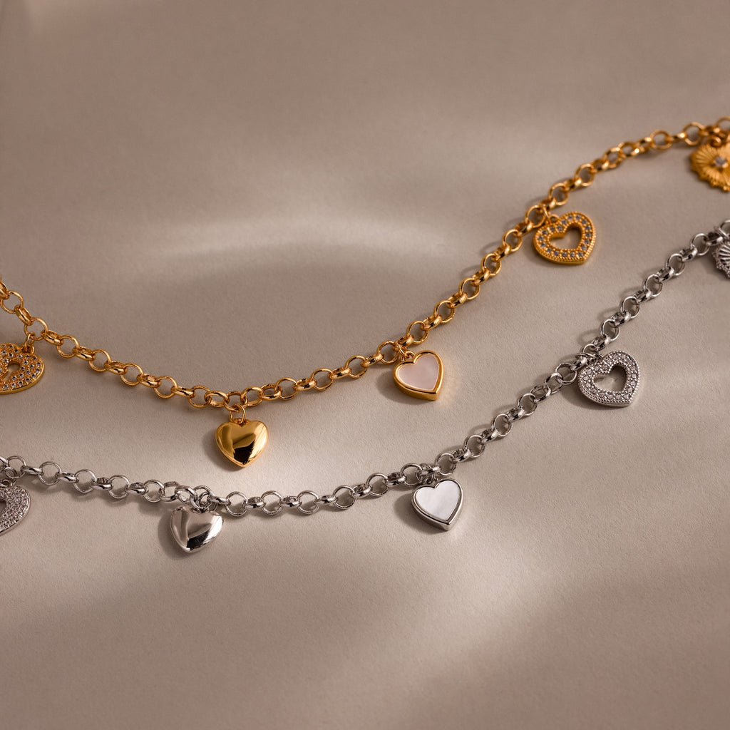 The Heart Charm Necklace in 18K Gold, featuring bold chain links and delicate heart charms, is displayed on a neutral beige background.