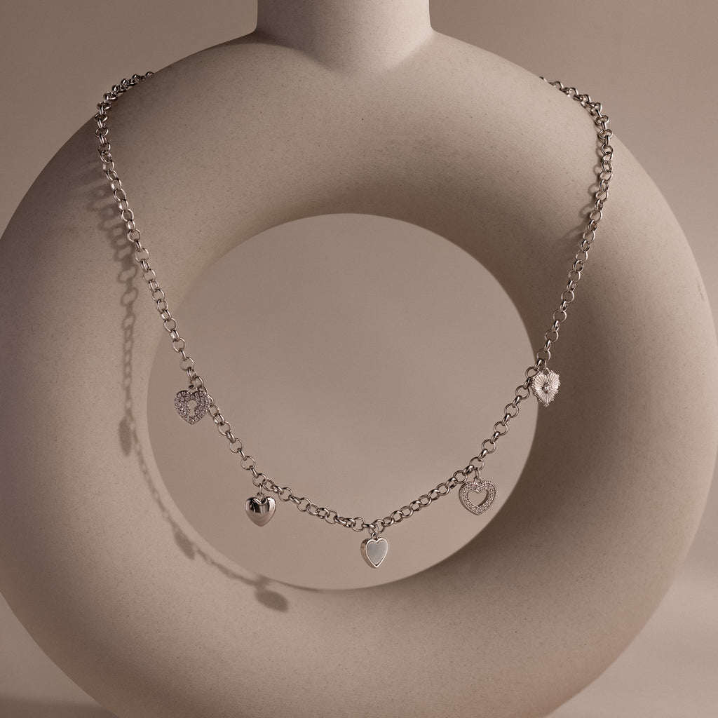 Silver heart charm necklace with five unique heart pendants displayed on a sculptural ceramic stand.