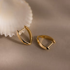 Gold leaf-shaped hoop earrings with a high-polish finish, displayed on a neutral beige surface beside a white scallop shell.