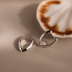 Pair of sculptural leaf-shaped silver hoop earrings displayed beside a white seashell with warm brown accents on a soft beige surface.