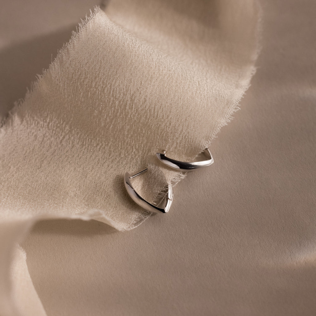 Pair of sculptural silver leaf-shaped hoop earrings displayed on a soft, textured ivory fabric with a neutral beige background.