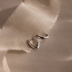 Pair of sculptural silver leaf-shaped hoop earrings displayed on a soft, textured ivory fabric with a neutral beige background.