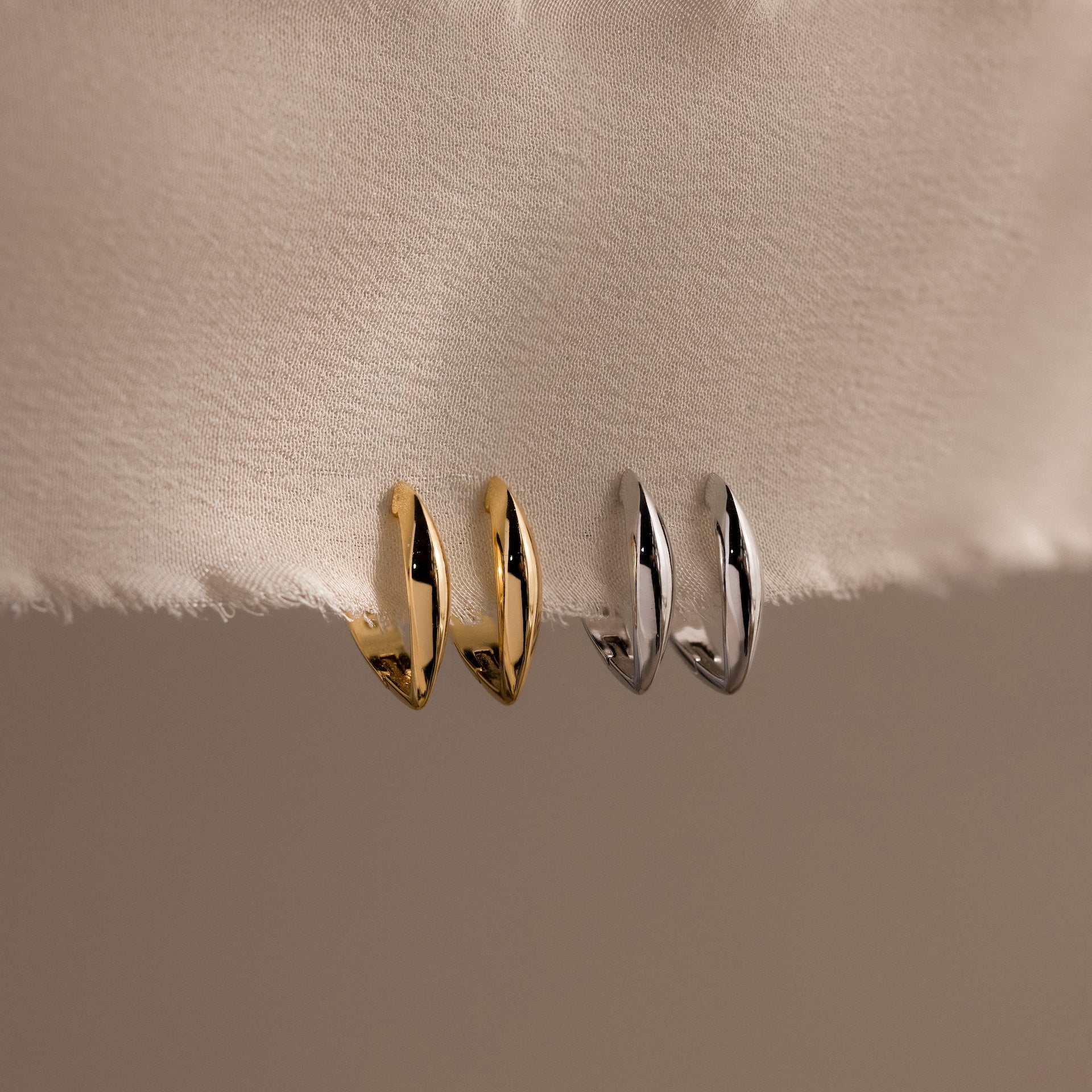 Four small sculptural leaf-shaped hoop earrings—two in polished gold and two in polished silver—displayed vertically hanging from a soft, textured cream fabric.