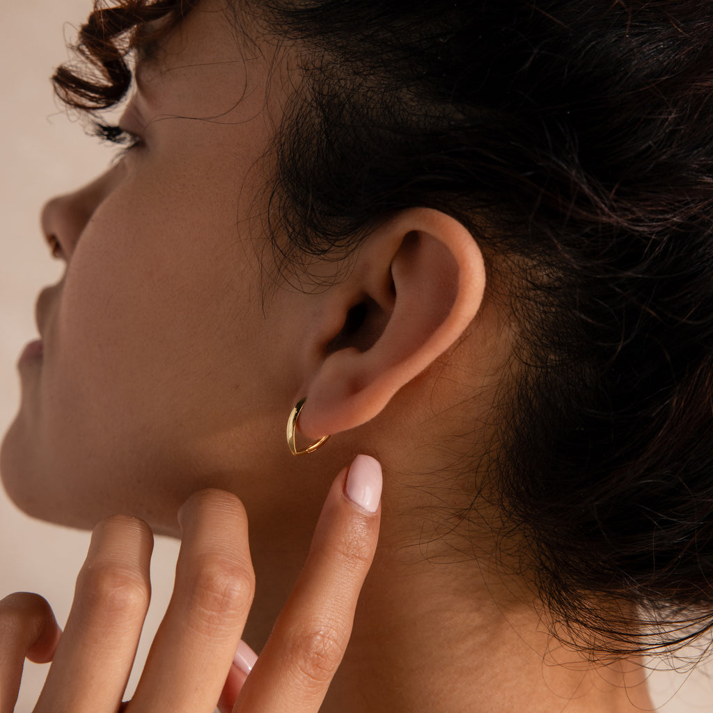 Model wearing a small, polished gold leaf-shaped hoop earring, gently touching her earlobe against a soft neutral background.