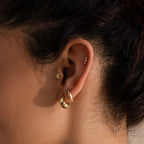 Close-up of a woman's ear adorned with multiple gold earrings, including a pair of chunky leaf-shaped hoops, a petite floral stud, and a curved cluster of tiny gemstones in the helix.