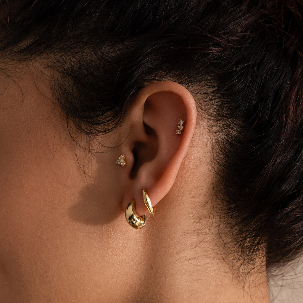 Close-up of a model's ear adorned with a gold oval huggie earring, chunky hoop, and two delicate pavé stud earrings.