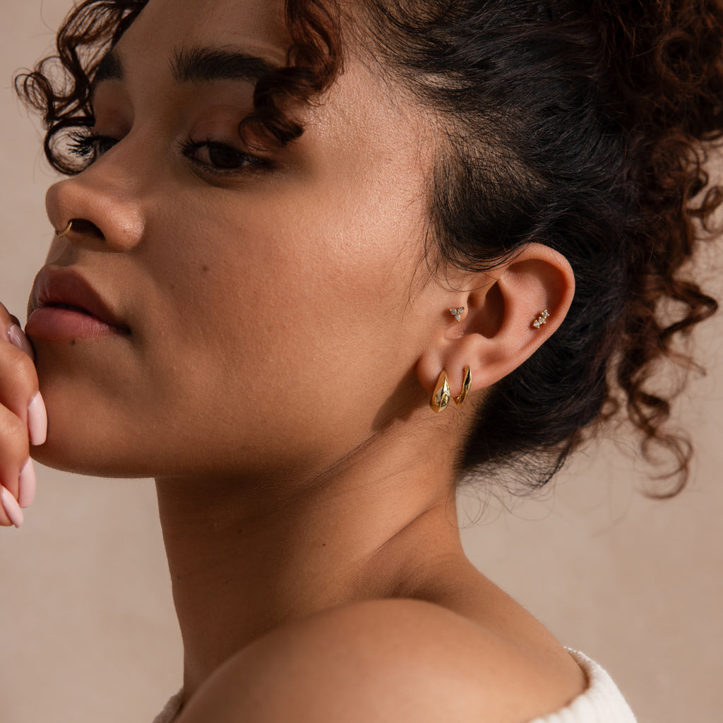 Model wearing two sculptural gold hoop earrings on the lobe, paired with dainty gemstone studs on the upper ear.