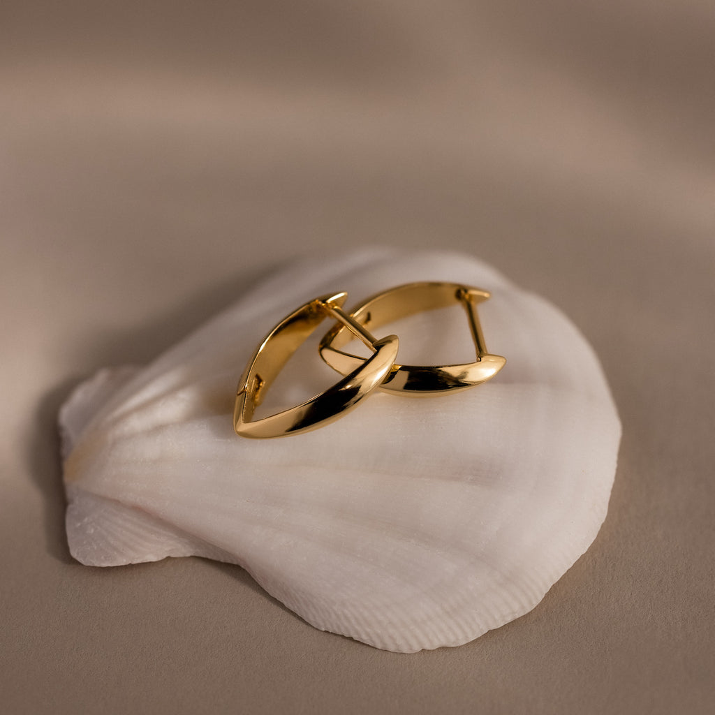 Pair of polished gold leaf-shaped hoop earrings with a sculptural, tapered design displayed on a white seashell against a soft beige background.