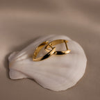 Pair of polished gold leaf-shaped hoop earrings with a sculptural, tapered design displayed on a white seashell against a soft beige background.