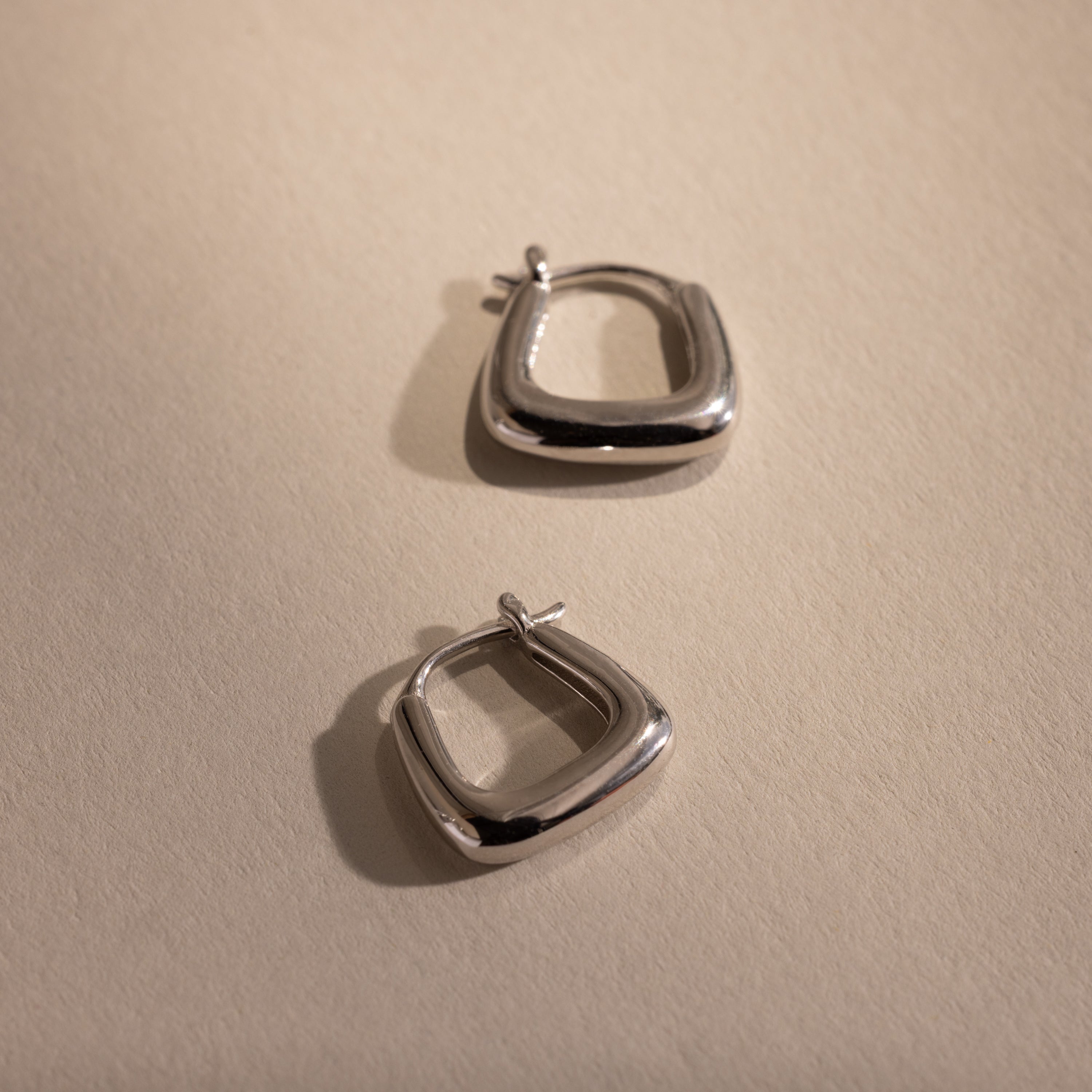 Pair of silver geometric hoop earrings displayed on a neutral background, highlighting their modern square shape and polished finish.