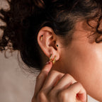 Close-up of ear with stacked gold piercings, featuring a bold geometric square gold hoop earring as the focal point, styled with textured hoops and black onyx studs.