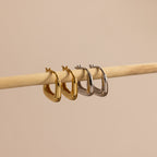 Gold and silver geometric square hoop earrings displayed side by side on a wooden rod, showcasing their bold modern shape and sleek finish.