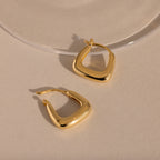Pair of gold geometric square hoop earrings resting on a neutral surface.