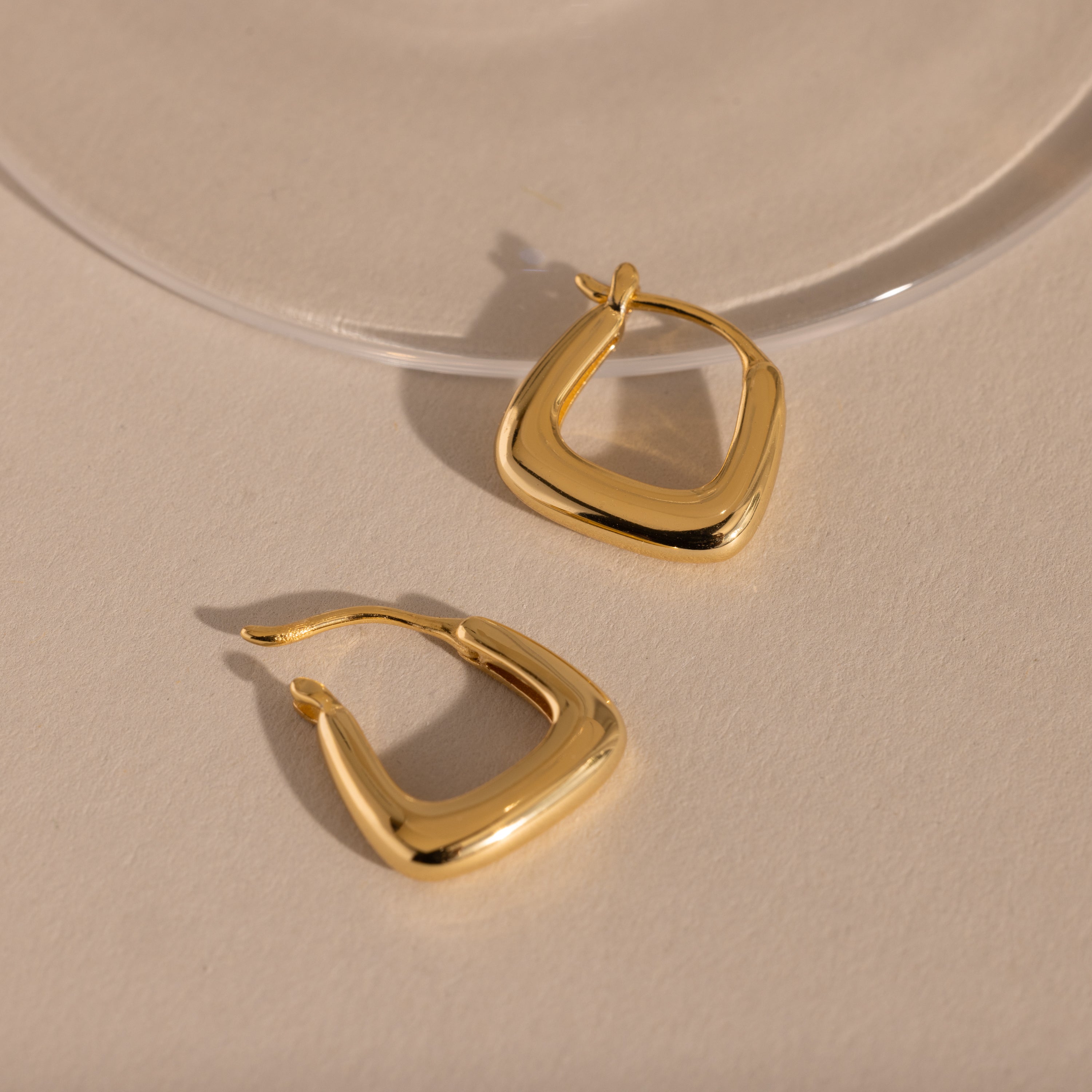 Pair of gold geometric square hoop earrings resting on a neutral surface.