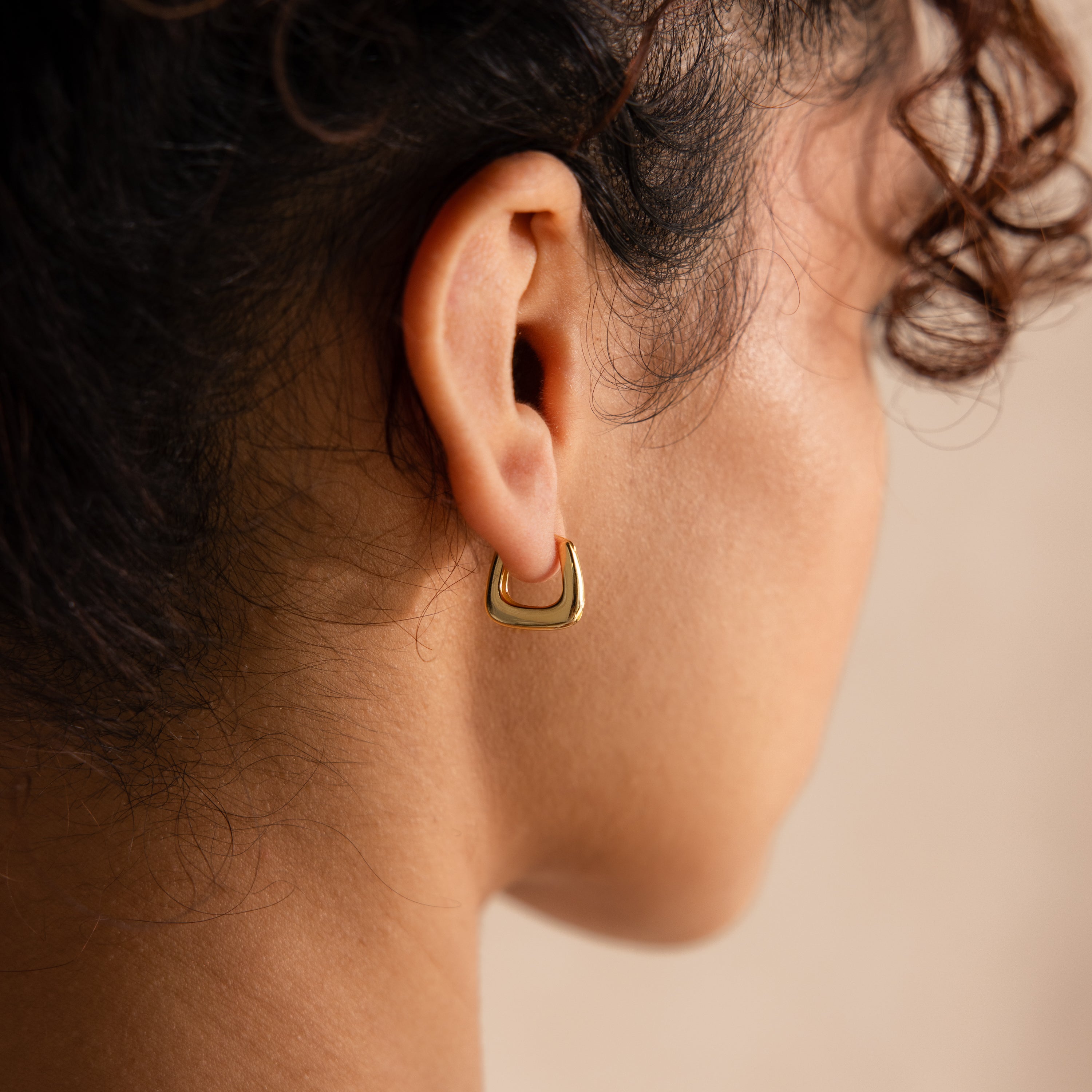 Close-up of gold geometric hoop earring worn on the ear, showcasing its bold, sculptural square shape against a soft neutral background.