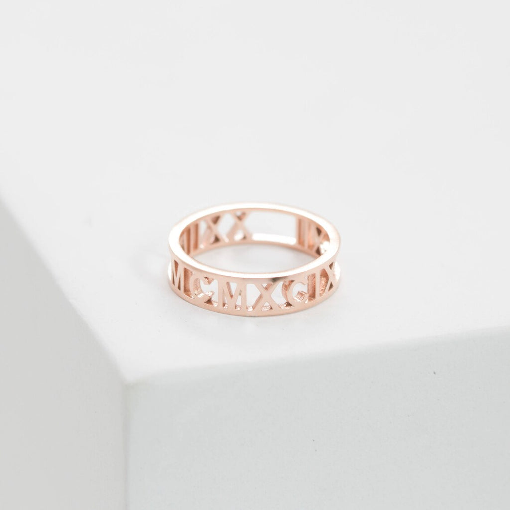 The Roman Numerals Ring in Rose Gold features elegant cut-out detailing, making it ideal for marking personal milestones or special dates. Shown on a white surface for a refined presentation.