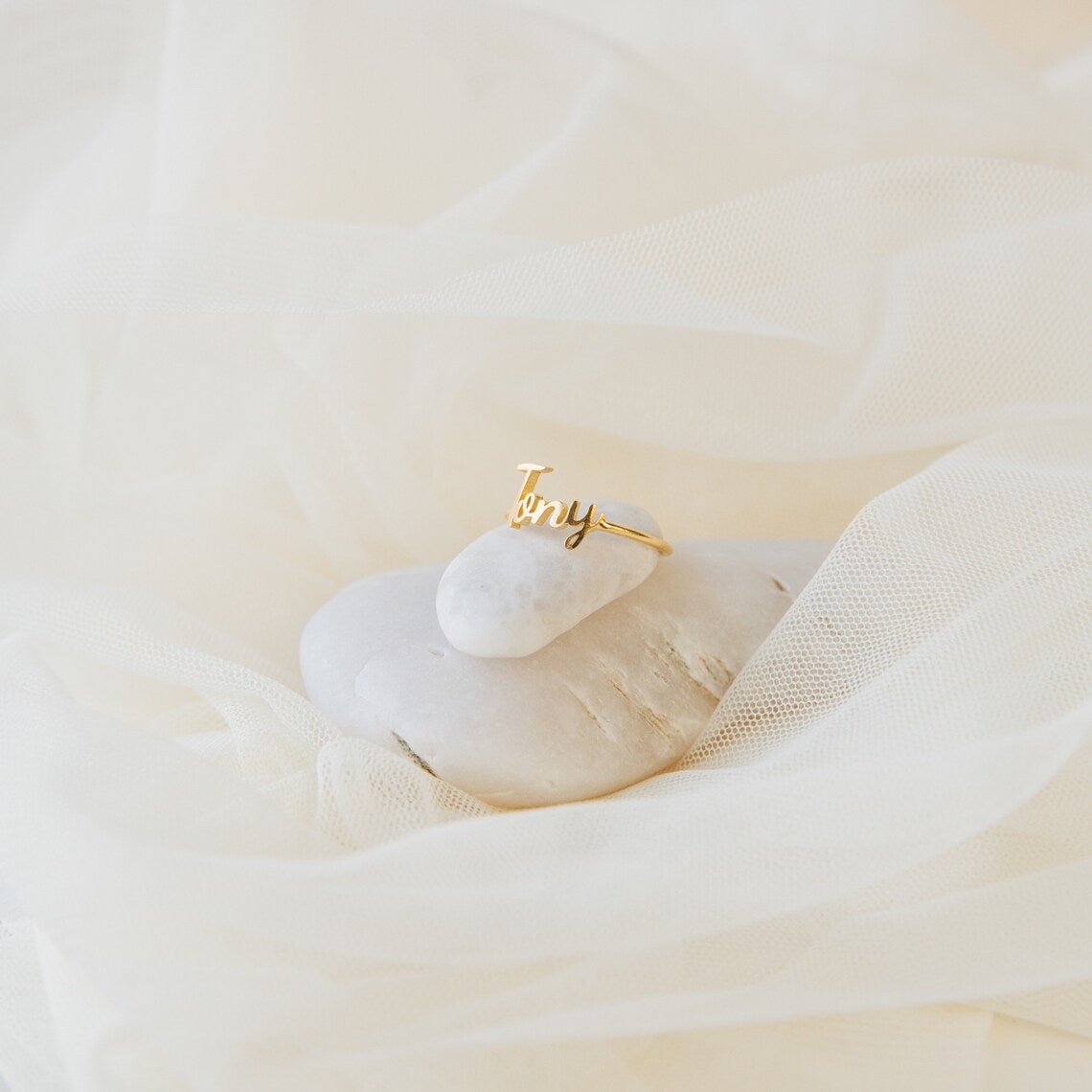The Grace Script Name Ring in 18K Gold, personalized with "Tony" in a script font, rests on a white stone and is surrounded by soft, sheer fabric.