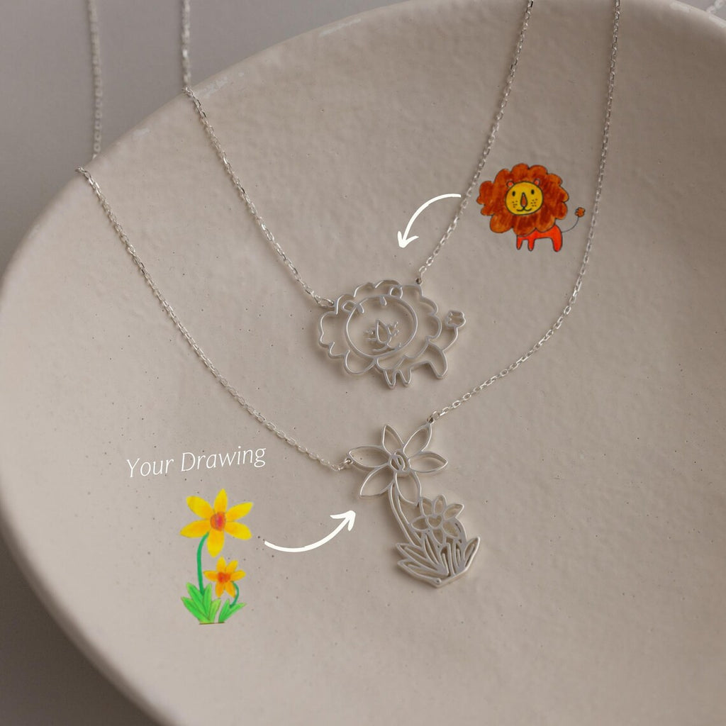 Two silver Kids Drawing Necklaces rest on a dish, each featuring a pendant recreated from a child’s drawing—a lion and a yellow flower—making these unique, personalized pieces of wearable art the perfect gift.