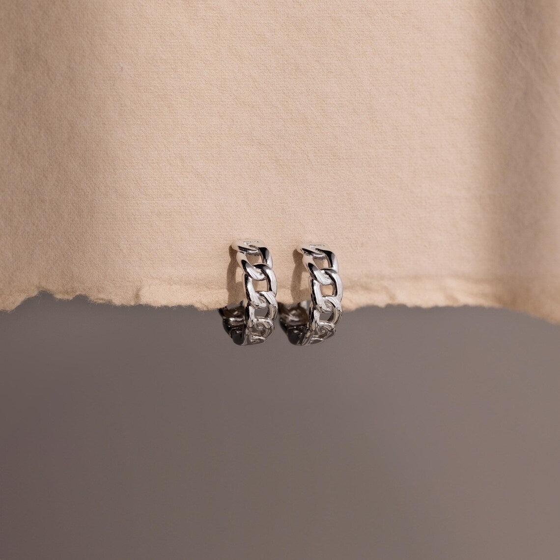 Pair of silver chain hoop earrings on a textured beige surface.