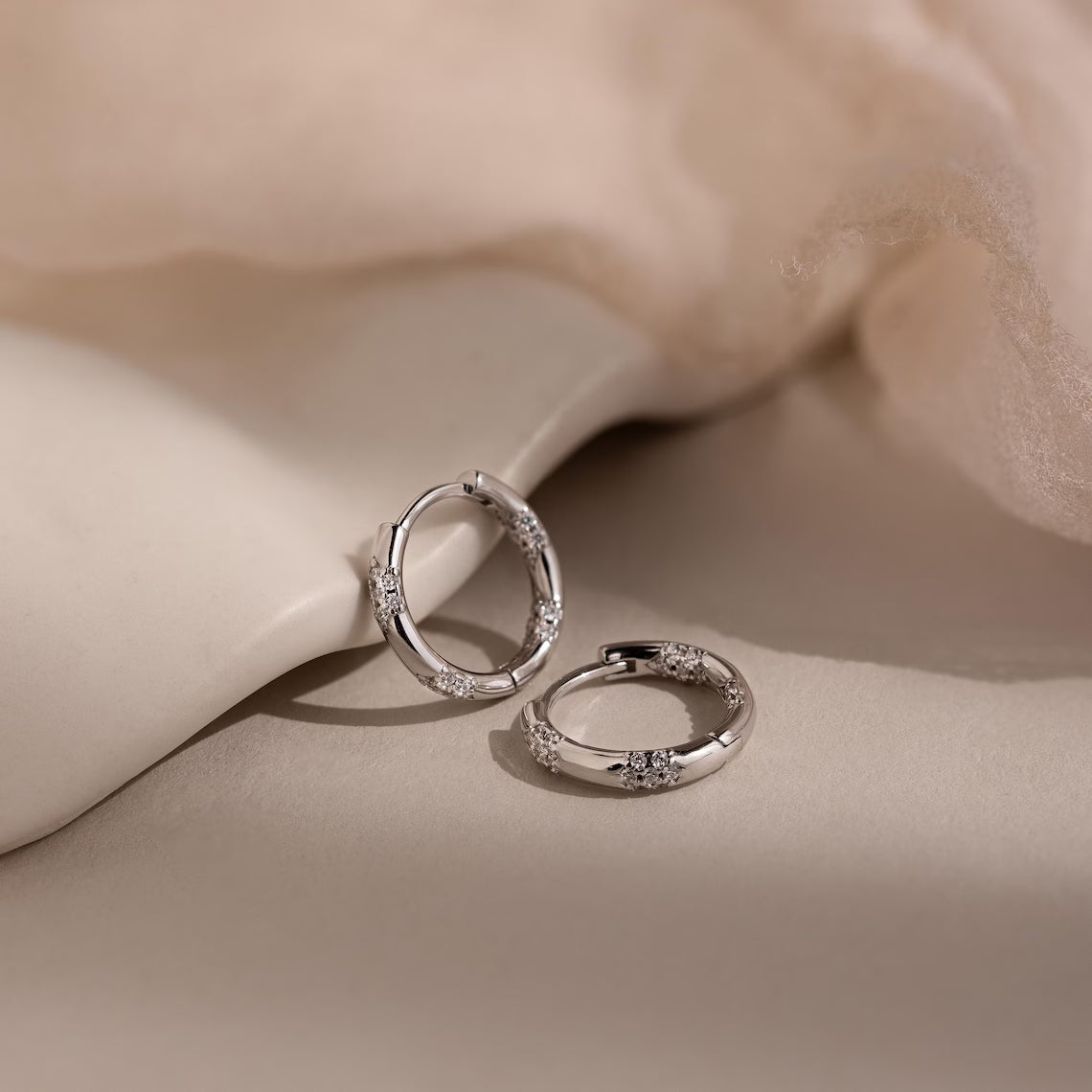 Silver hoop earrings with small diamonds on a beige fabric background