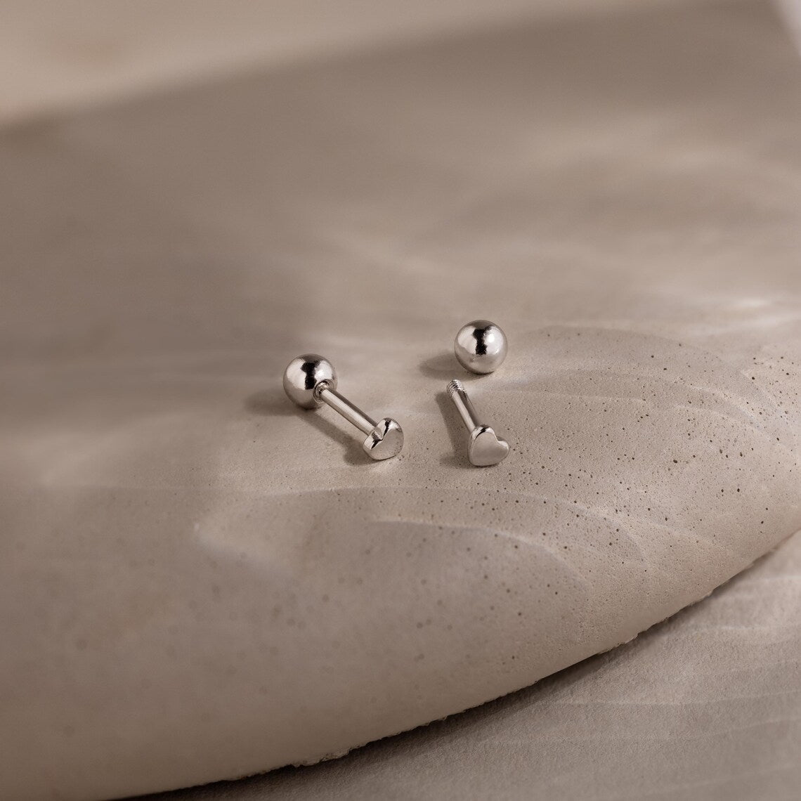 Two dainty silver heart earrings on a beige stone surface
