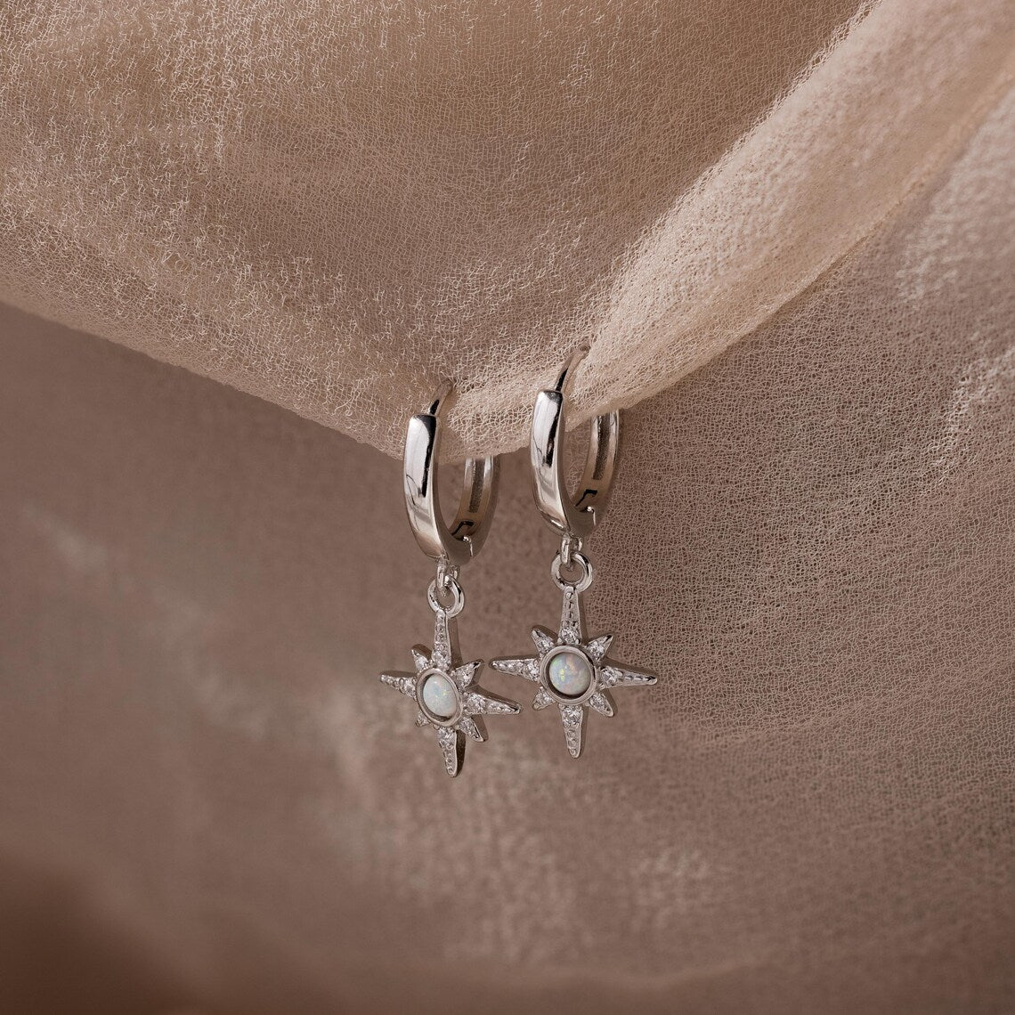 Pair of hanging silver opal hoop earrings with star designs on a beige fabric background