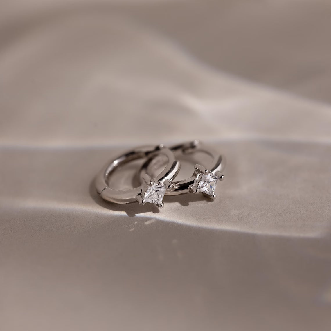 Silver huggie earrings with two diamond stones on a beige background