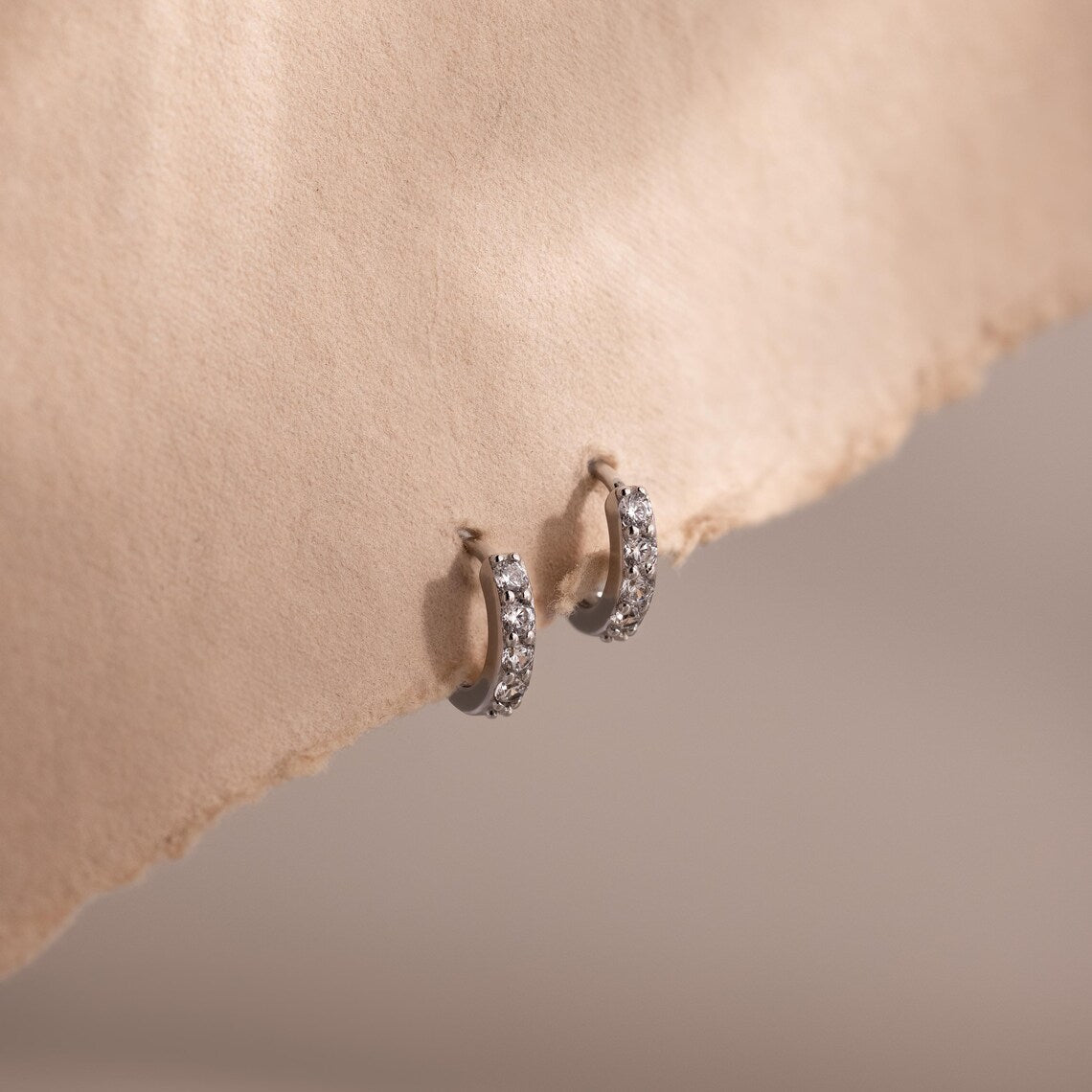 Pair of silver clear gemstone hoop earrings on a beige fabric background