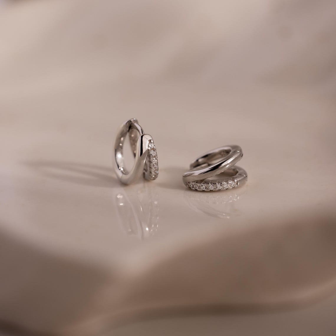 Two silver diamond hoop earrings on a beige surface