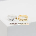A silver Handwriting Band engraved with "I love you" and a gold custom ring with "miss you right now" rest on a white surface—ideal as heartfelt Christmas gifts.