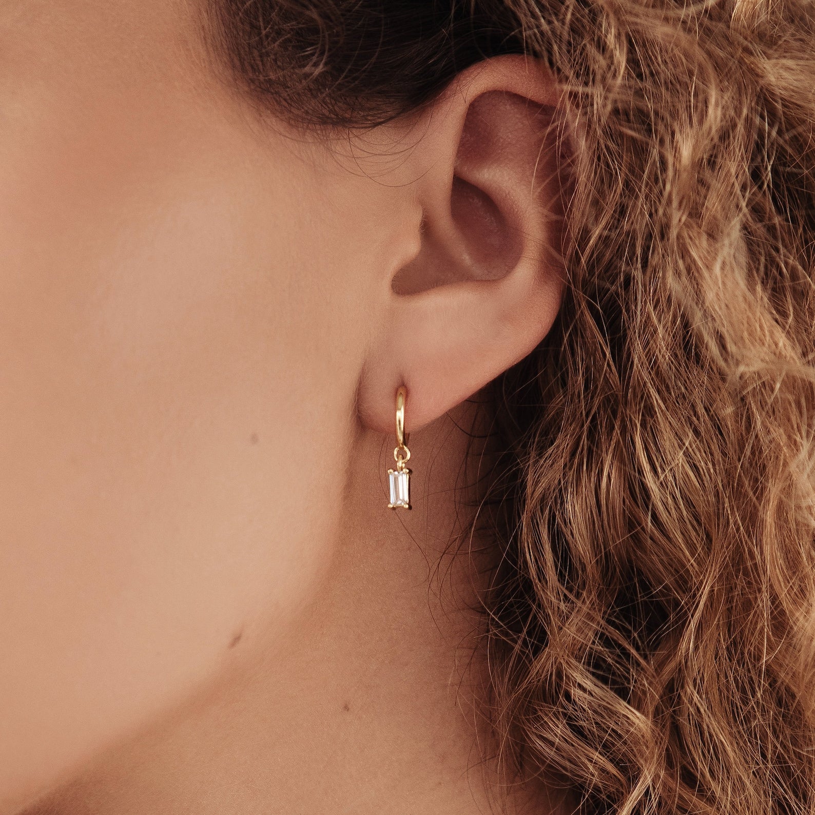 Close-up of a woman’s ear wearing Lys Baguette Hoops—gold earrings designed with classic elegance and a rectangular clear gemstone pendant.