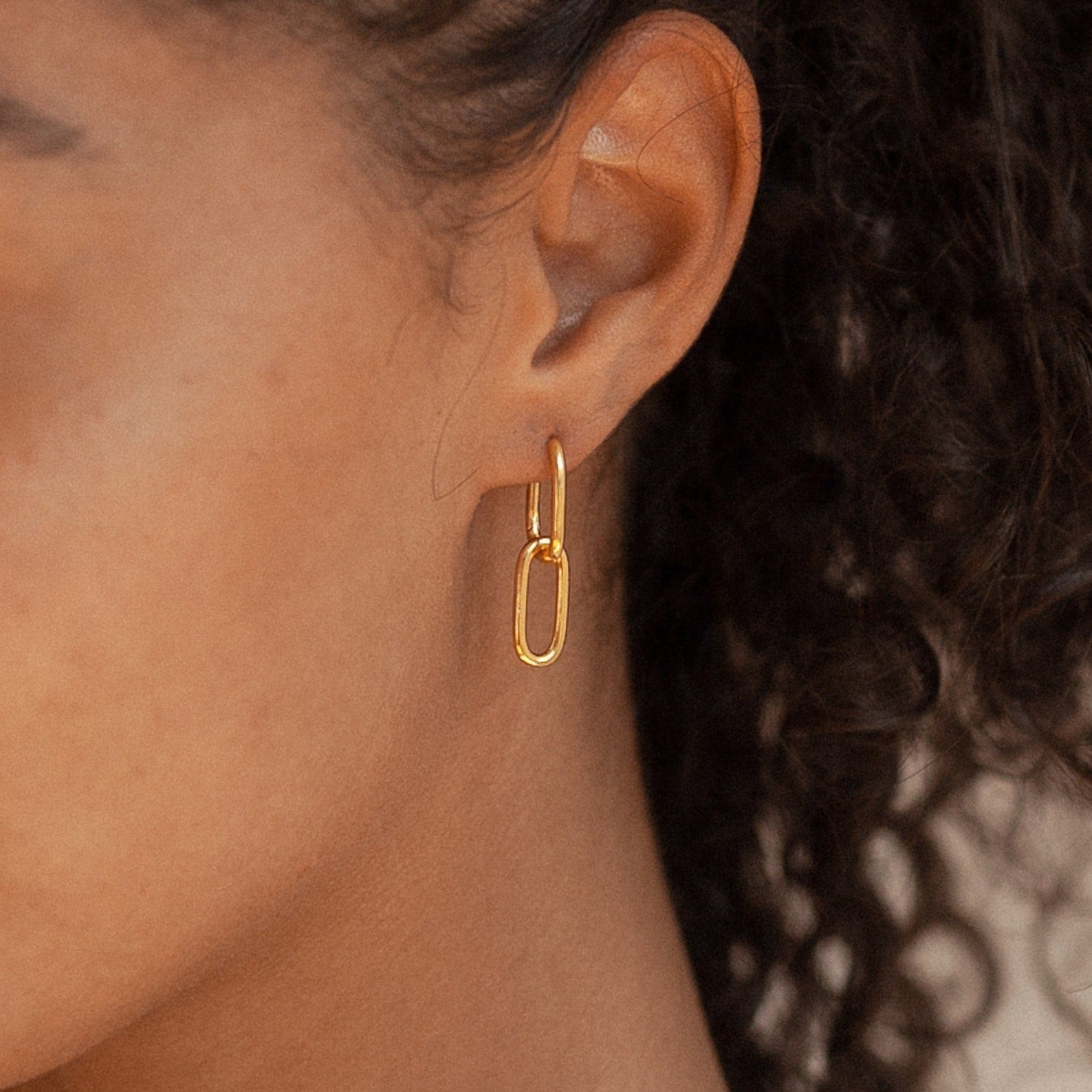 Close-up of a person wearing a gold oval-shaped hoop earring from the Linked Necklace & Earrings Set, with curly hair in the background.