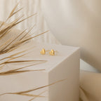 Gold heart-shaped stud earrings on a white pedestal with dried grasses.
