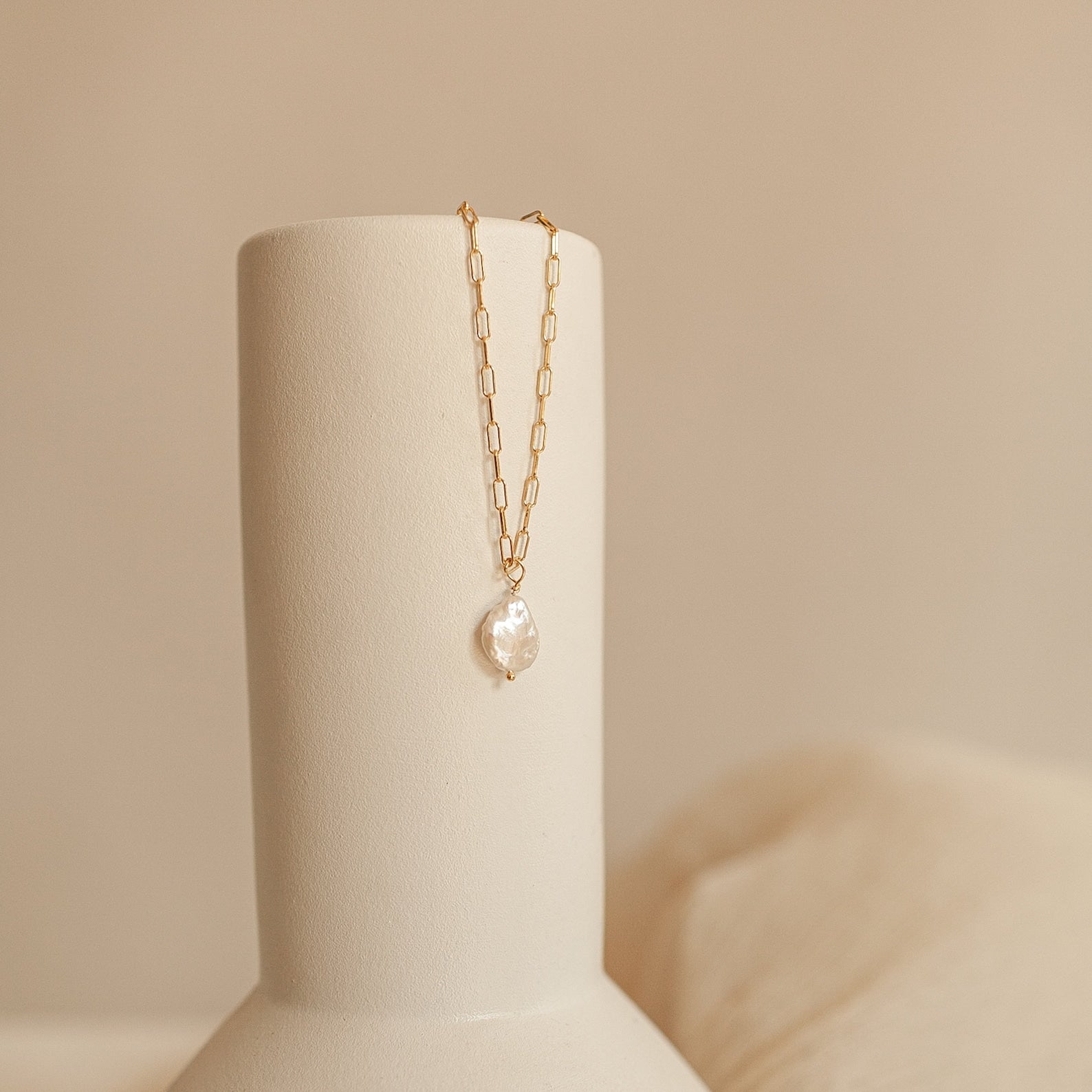 Gold paperclip chain necklace with a pearl pendant on a beige stand against a beige background