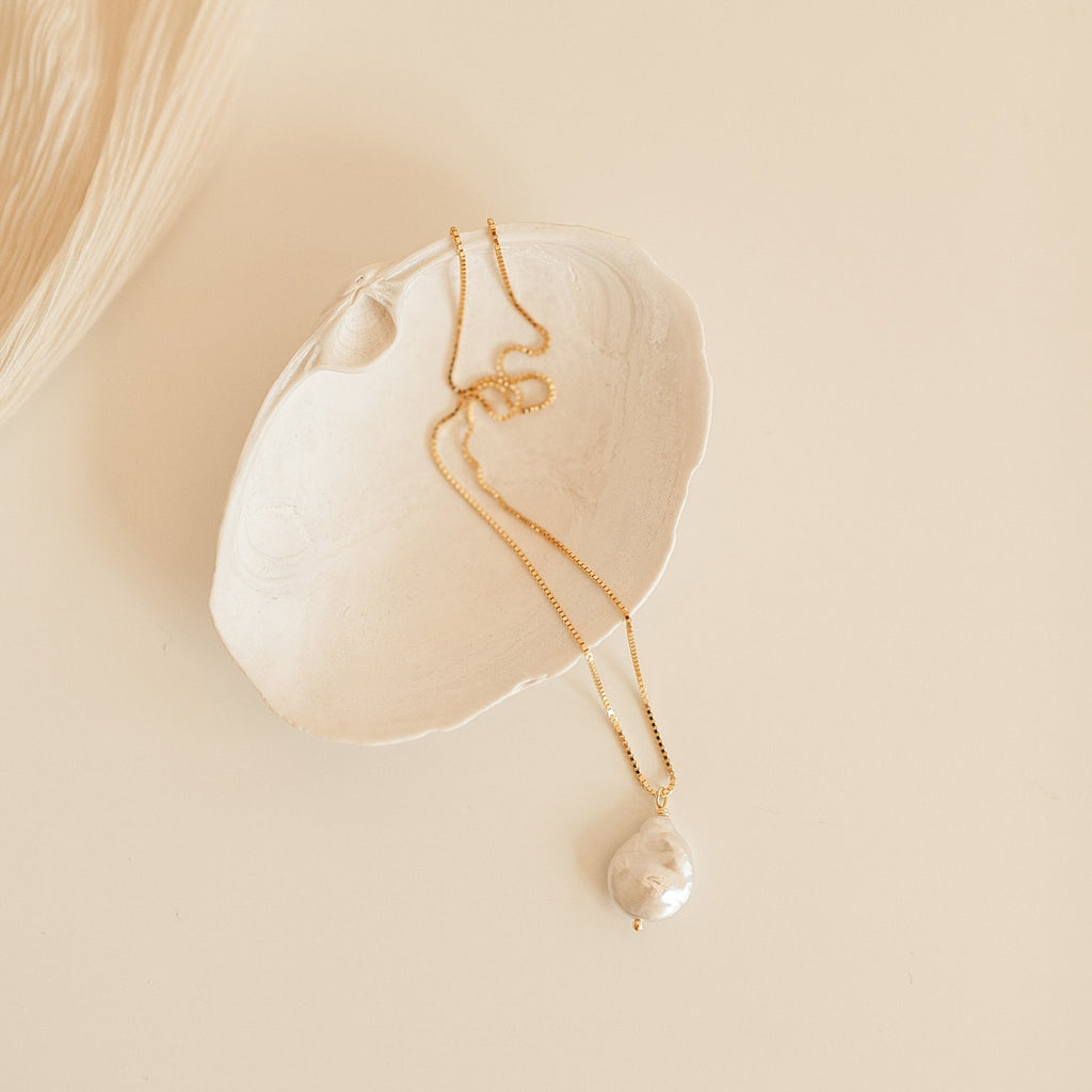 The Sadie Pearl Necklace is a gold baroque chain with a large freshwater pearl pendant, shown in a white seashell on beige—an elegant bridesmaid gift.