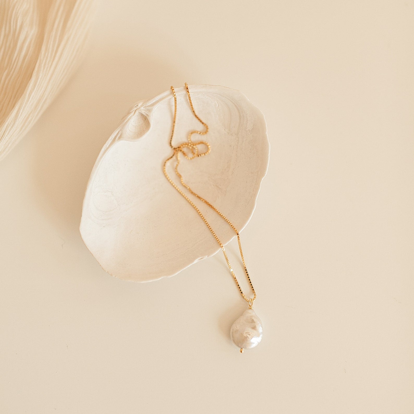 The Sadie Pearl Necklace is a gold baroque chain with a large freshwater pearl pendant, shown in a white seashell on beige—an elegant bridesmaid gift.