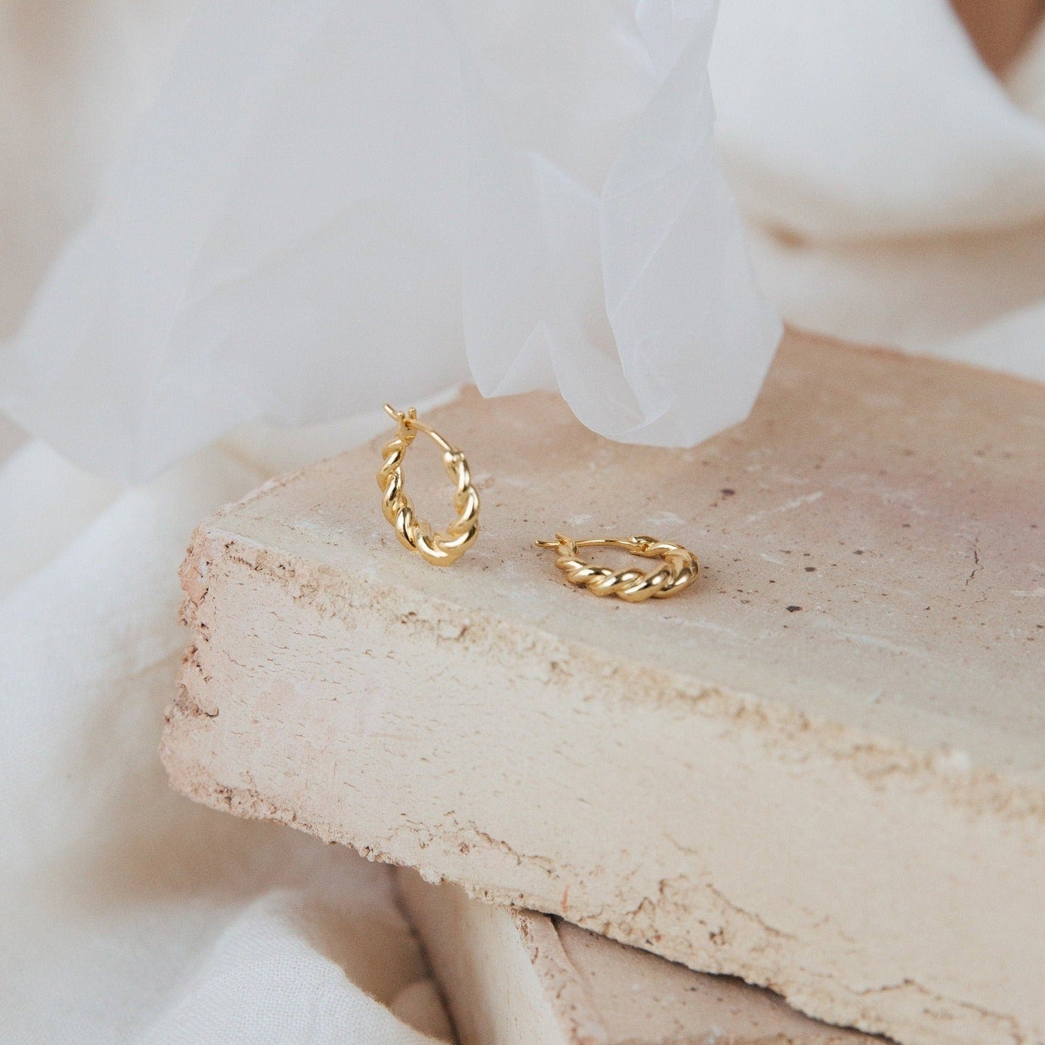 Naomi Twisted Hoops in 18K Gold displayed on light-colored stone slabs with a soft fabric background.