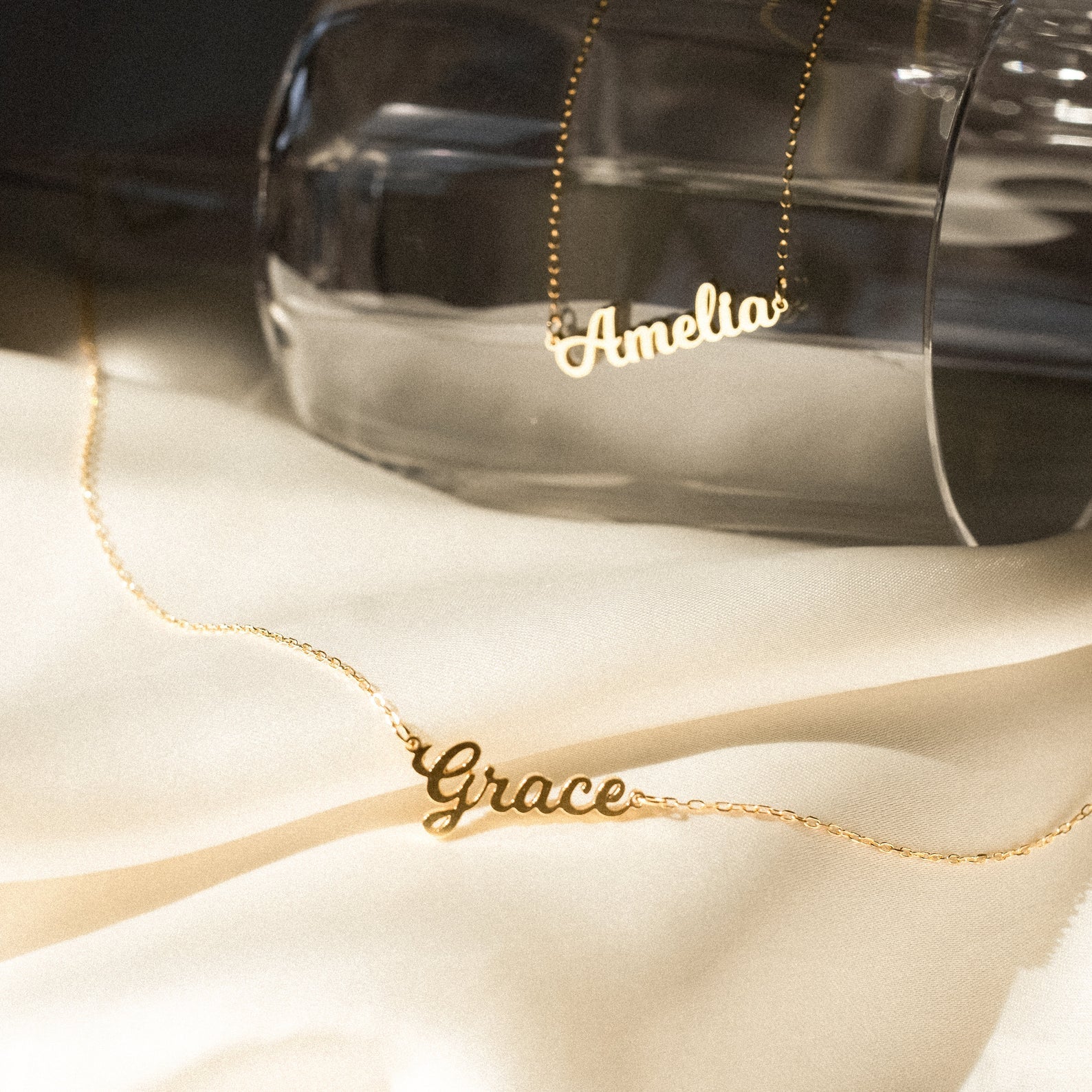 Two Mellow Name Necklaces, each personalized with the names Amelia and Grace, are displayed on a light fabric background.