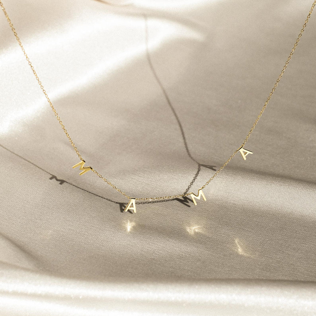 The Mama Letter Necklace features charms spelling MAMA, beautifully displayed on a soft, light fabric—an elegant tribute to motherhood and a thoughtful gift for moms.