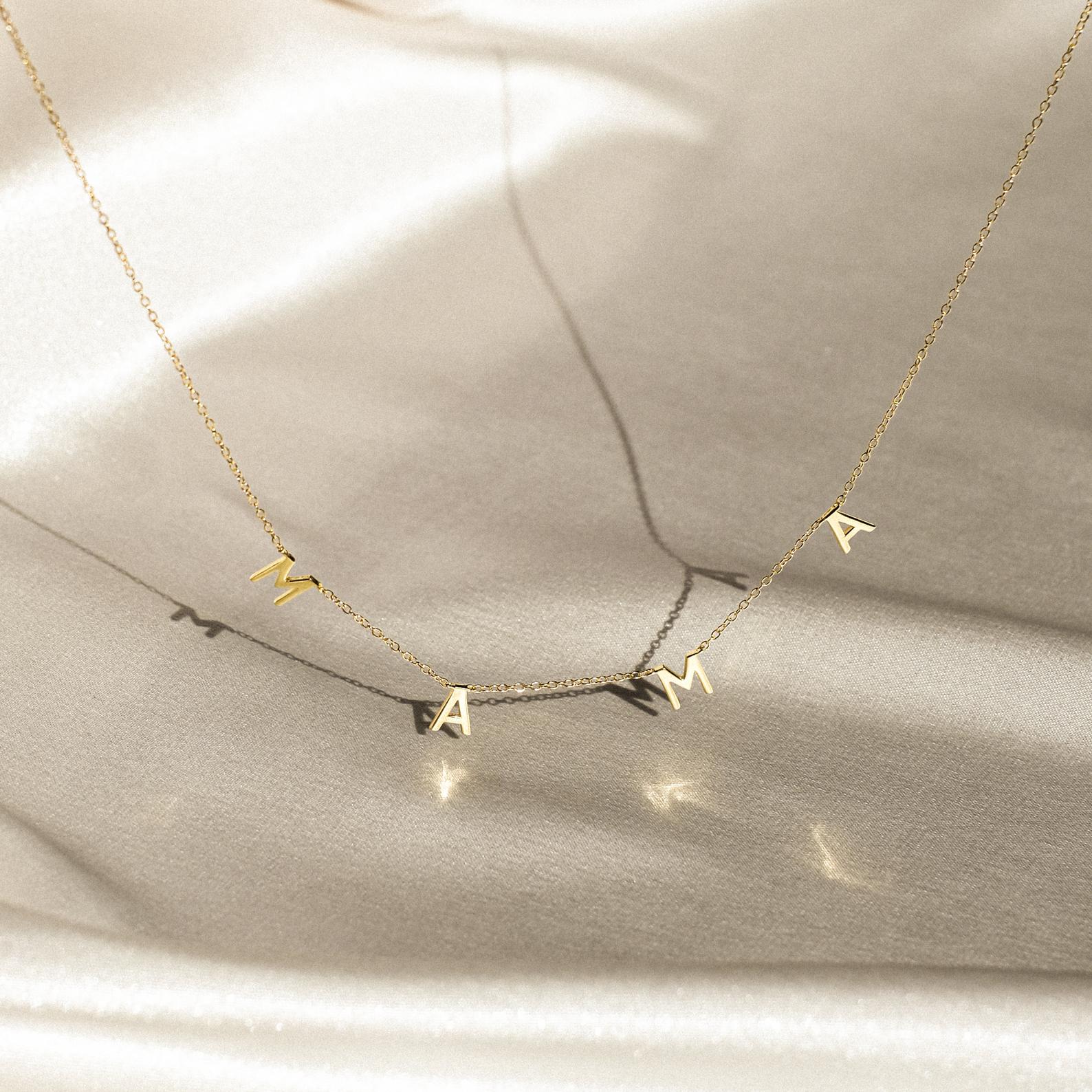 The Mama Letter Necklace features charms spelling MAMA, beautifully displayed on a soft, light fabric—an elegant tribute to motherhood and a thoughtful gift for moms.