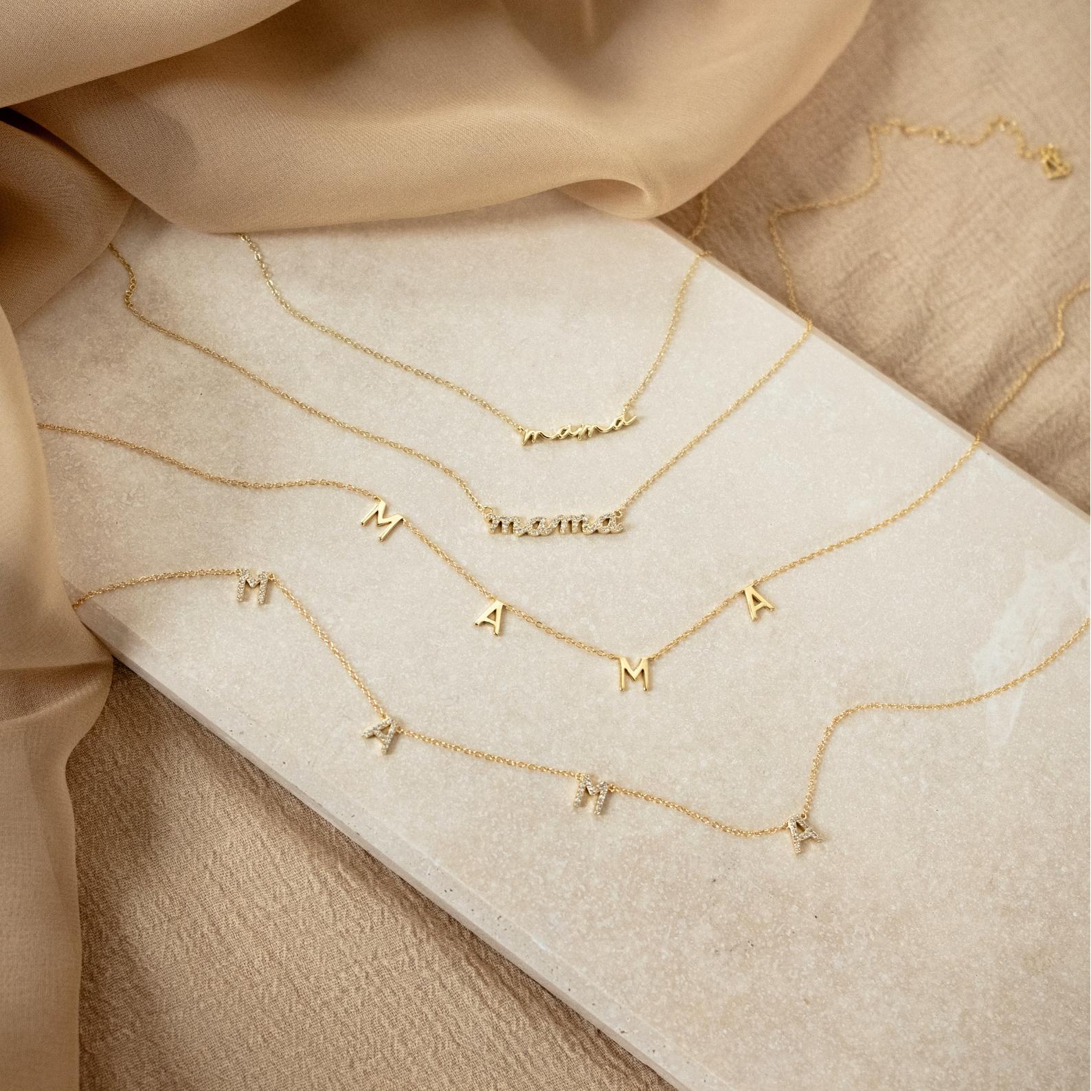 Four Mama Letter Necklaces rest on a beige surface among soft tan and beige fabric—an ideal mother gift and a lovely way to celebrate motherhood.