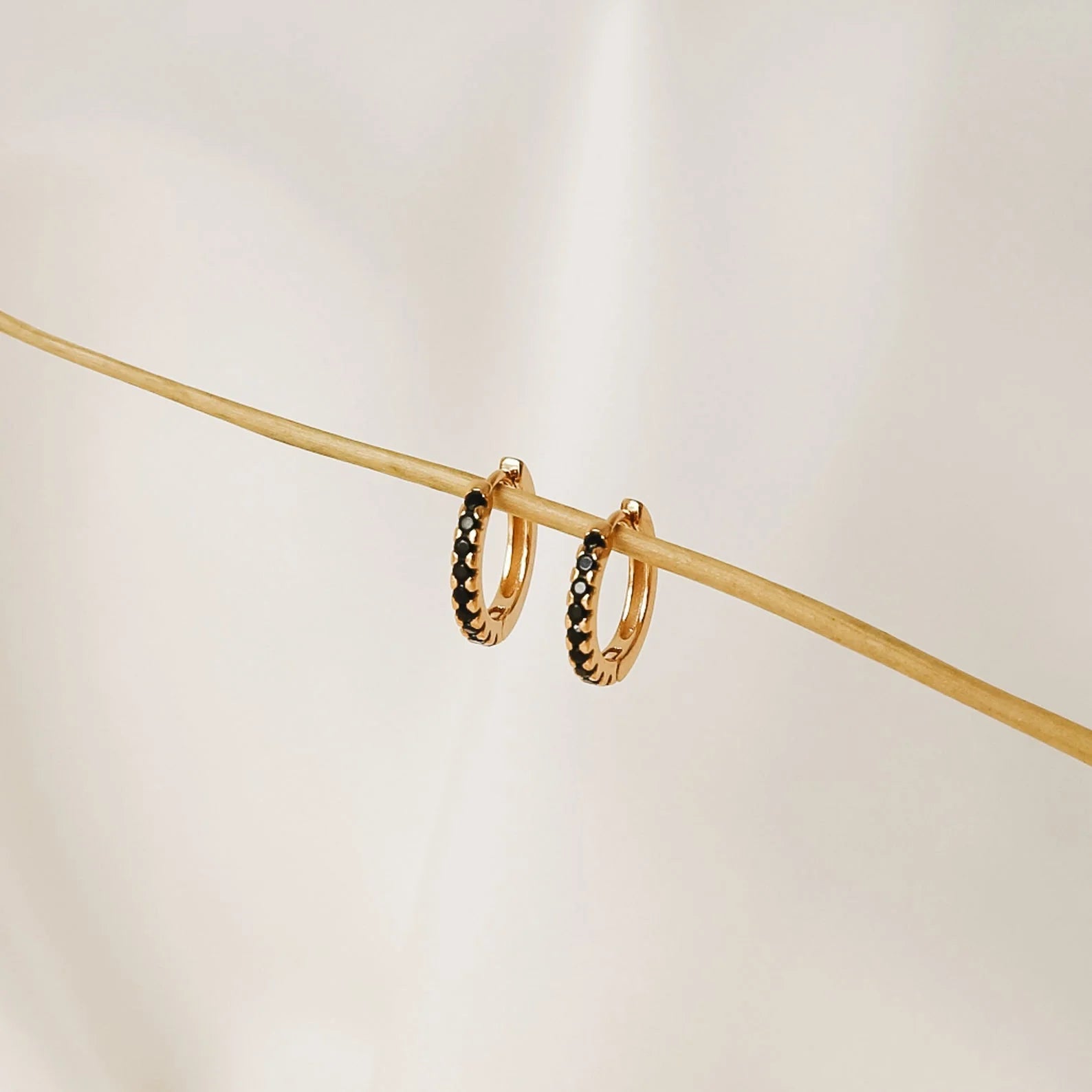 Onyx Diamond Earrings Set, gold hoops adorned with small black stones, hang on a thin beige stick against a soft cream background.