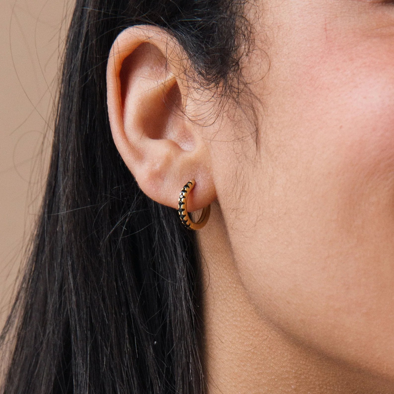 Close-up of a person’s ear wearing a small gold hoop earring with black stone accents, from the Onyx Diamond Earrings Set.