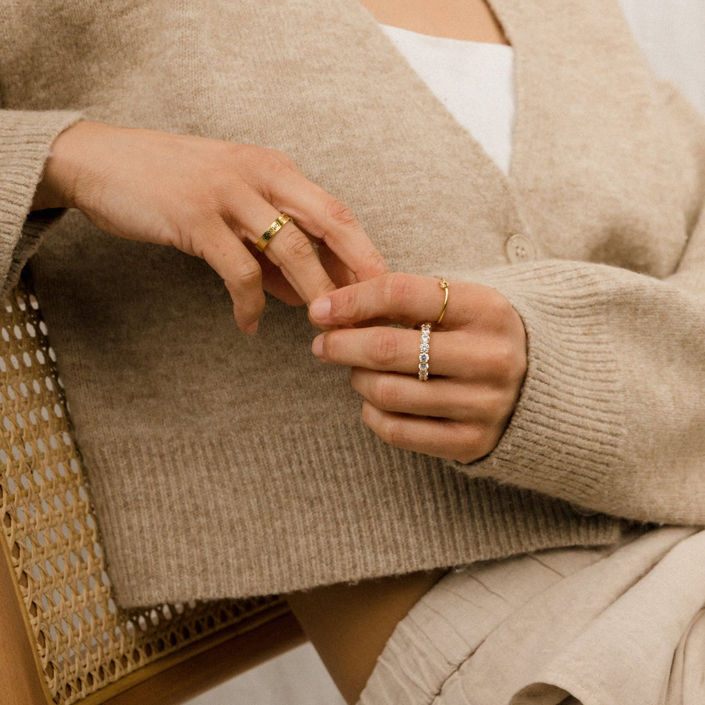 A woman in a beige cardigan sits on a woven chair, wearing the Knot Ring in 18K Gold on her fingers—a perfect symbolic gesture or thoughtful wedding gift.