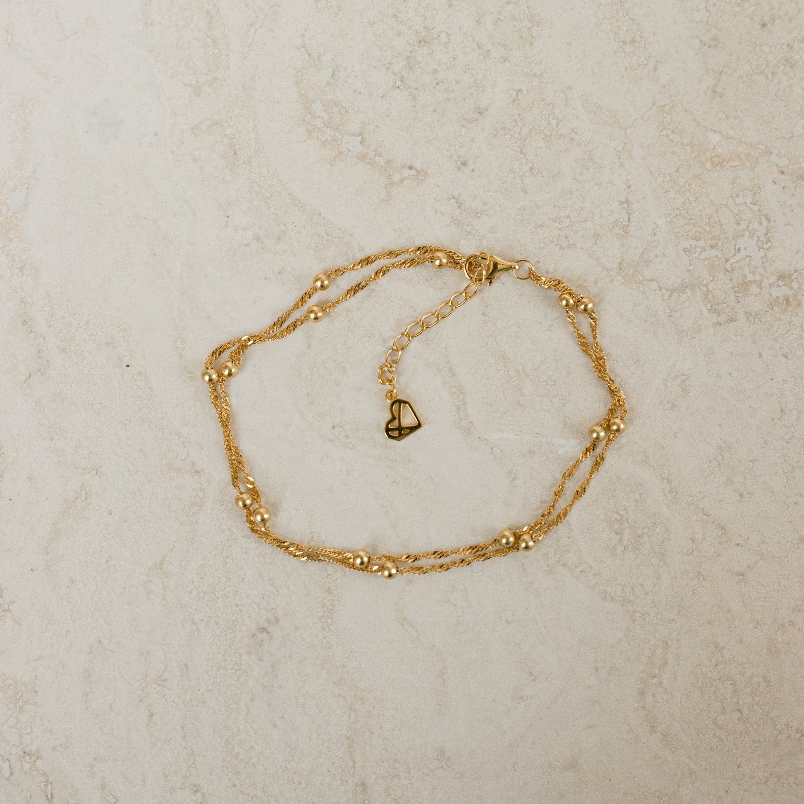 The Duo Bead Chain Bracelet, featuring a delicate gold chain with small beads and a heart-shaped clasp, rests on light marble—perfect to pair with your favorite necklace for a chic, layered look.