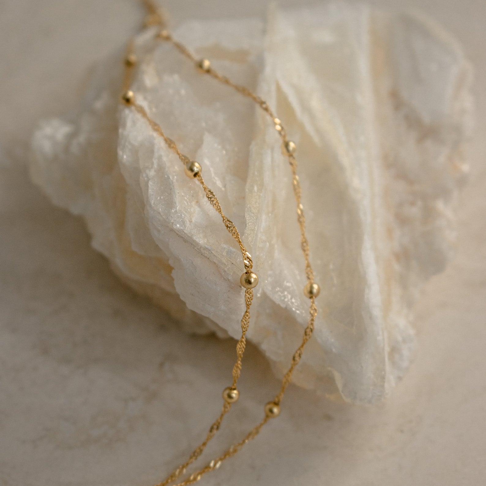 The Duo Bead Chain Anklet in Sterling Silver, featuring delicate small beads, is draped over a white crystal stone on a neutral background.