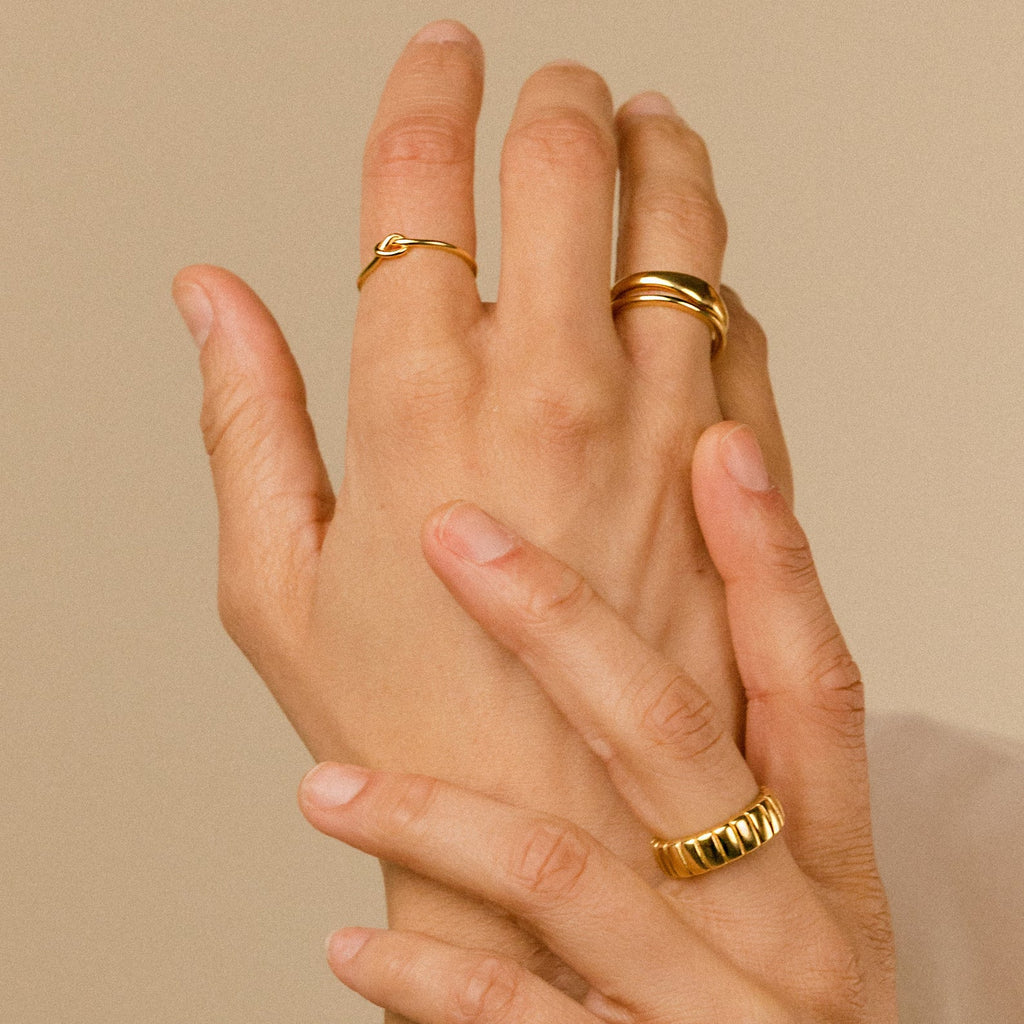 Two hands adorned with gold rings, featuring the Knot Ring, gently touch against a neutral background—a symbolic gesture of connection and love.