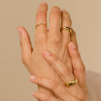 Two hands adorned with gold rings, featuring the Knot Ring, gently touch against a neutral background—a symbolic gesture of connection and love.