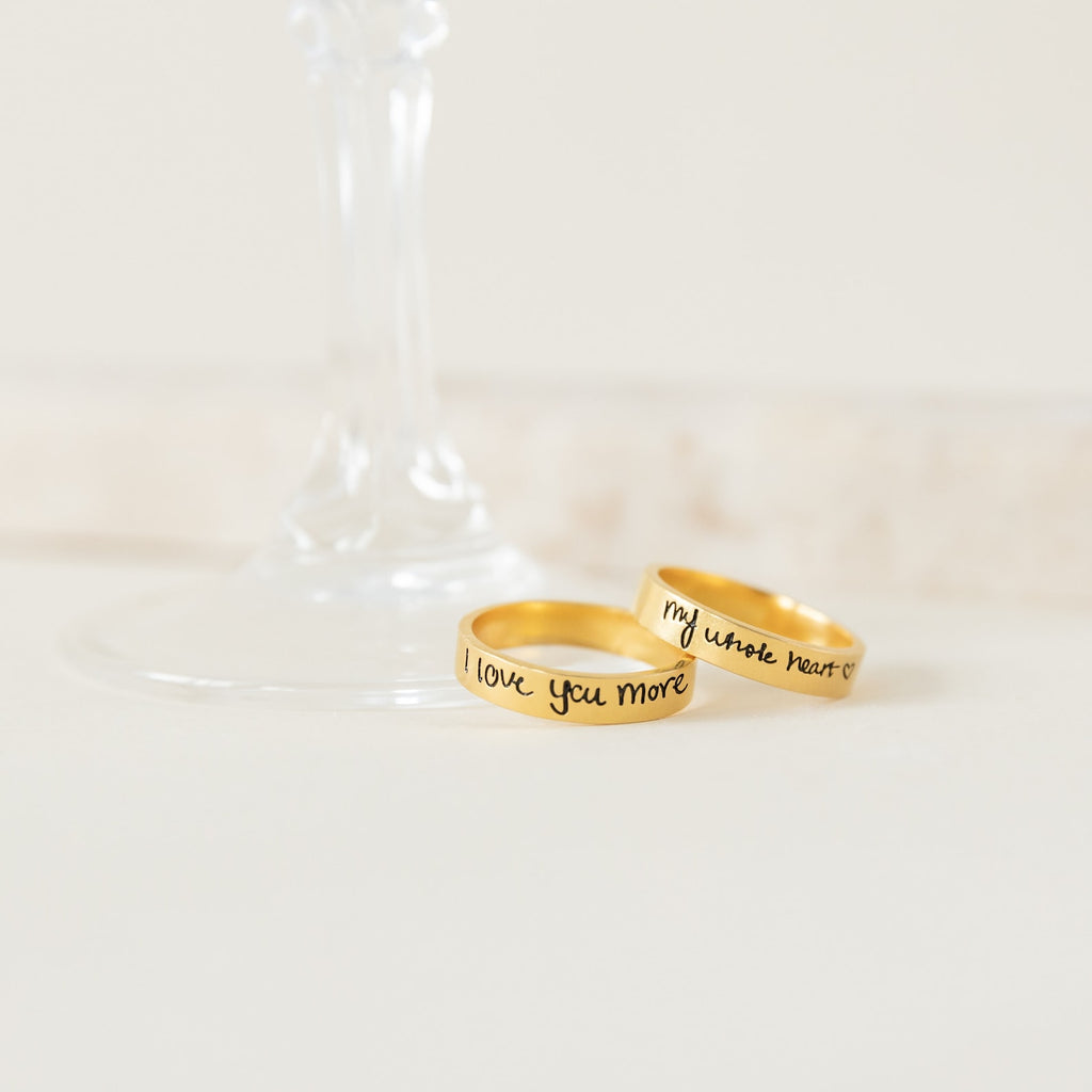 Two gold Handwriting Bands, engraved with "I love you more" and "my whole heart," rest by a wine glass base—ideal as a Christmas gift or personalized jewelry for someone special.